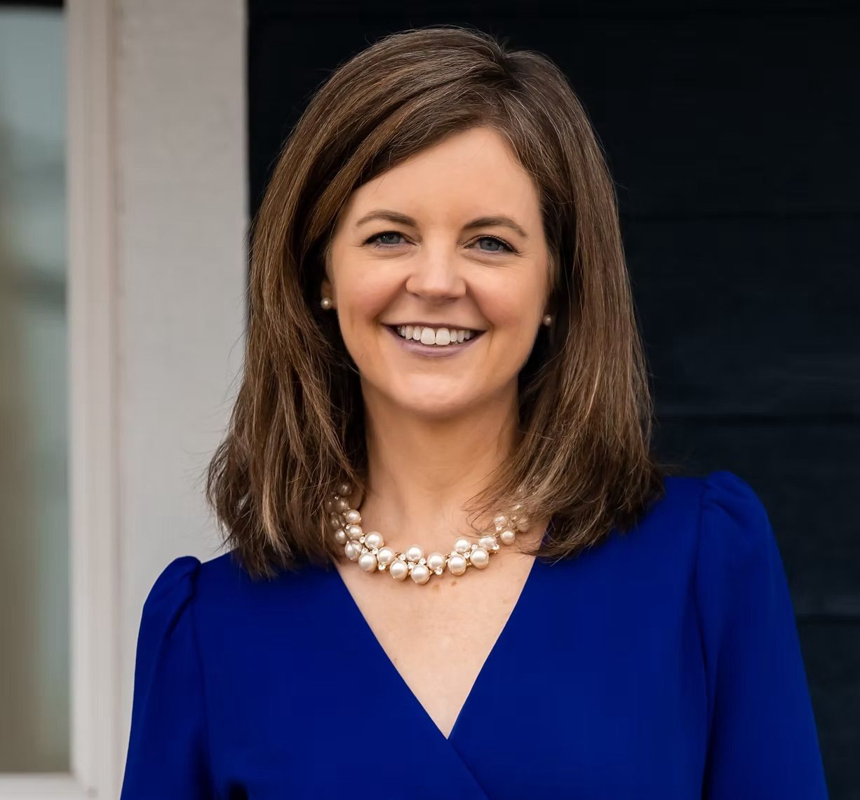 A woman wearing a blue dress and a pearl necklace is smiling.