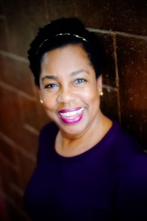 A woman in a purple shirt is smiling in front of a brick wall