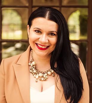 A woman wearing a tan jacket and a pearl necklace is smiling for the camera.