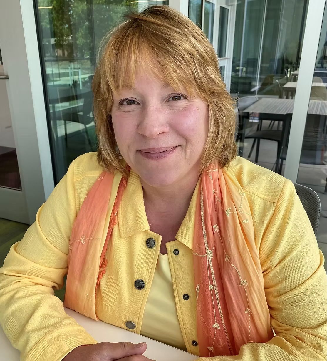 A woman wearing a yellow sweater and a scarf is sitting at a table.