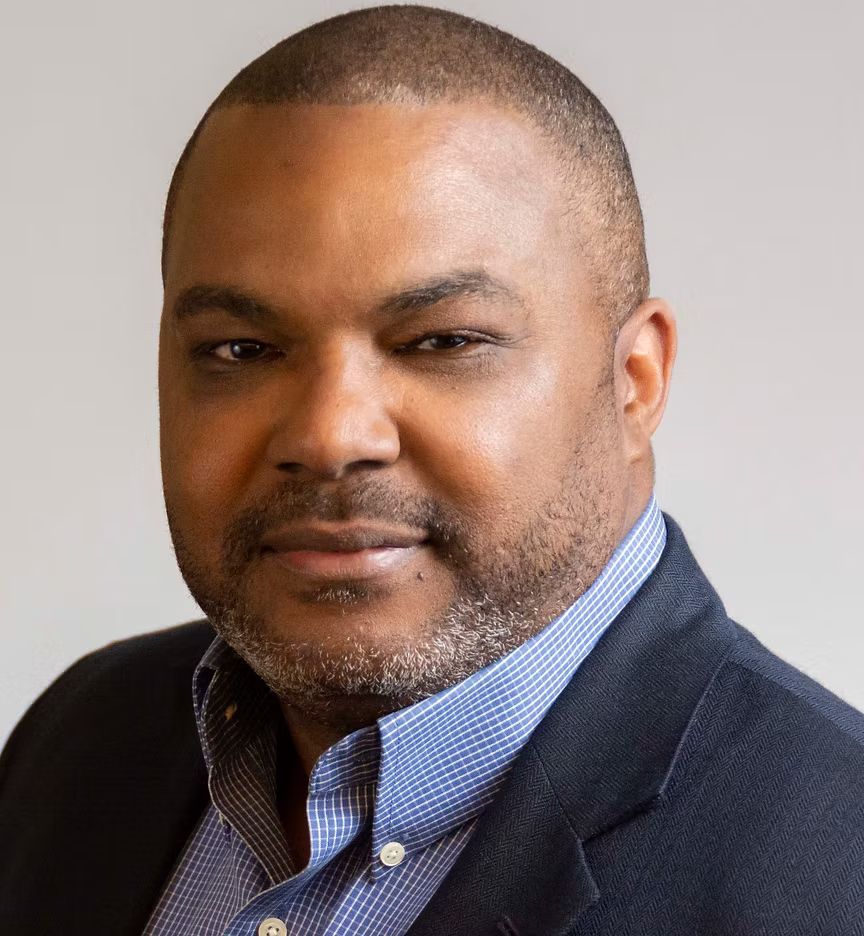 A man with a beard is wearing a suit and a blue and white checkered shirt.