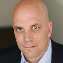 A bald man in a suit and blue shirt is looking at the camera.