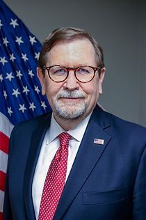 A man in a suit and tie is standing in front of an american flag.