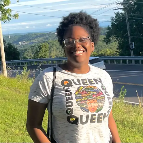 A woman wearing glasses and a queen t-shirt is standing in front of a road.