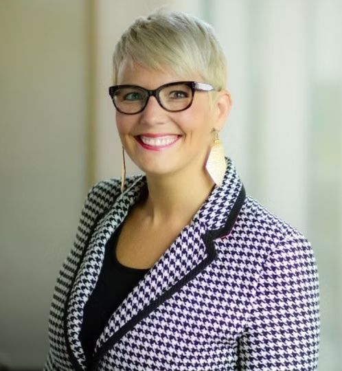 A woman wearing glasses and a black and white jacket is smiling.
