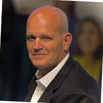 A bald man in a suit and white shirt is looking at the camera.