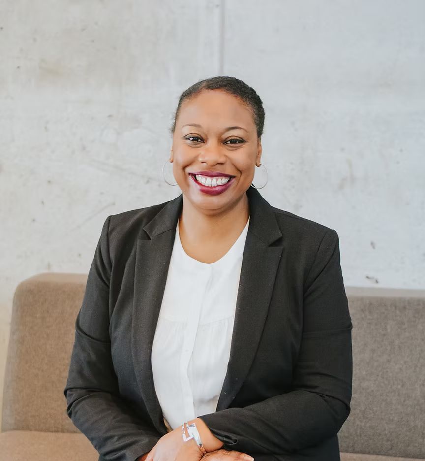 A woman in a black jacket and white shirt is smiling while sitting on a couch.