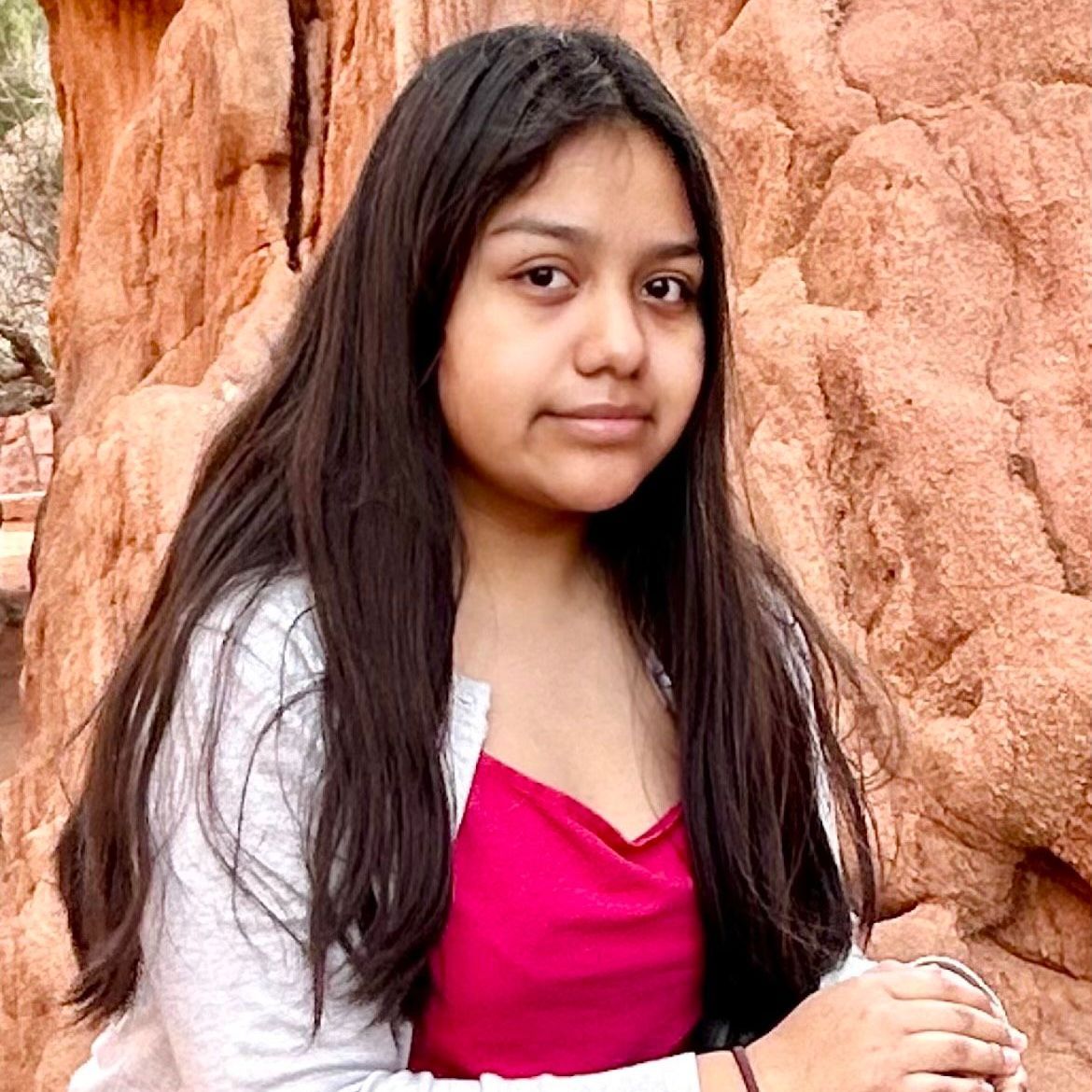 A young girl with long hair is standing in front of a rock formation.