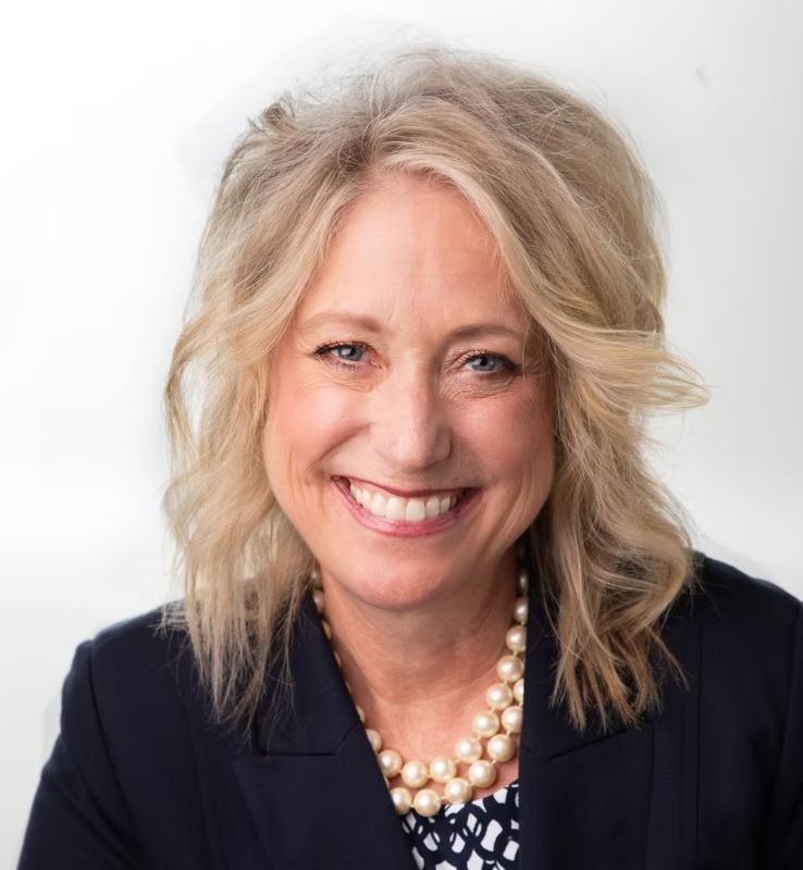 A woman wearing a blue jacket and a pearl necklace is smiling for the camera.
