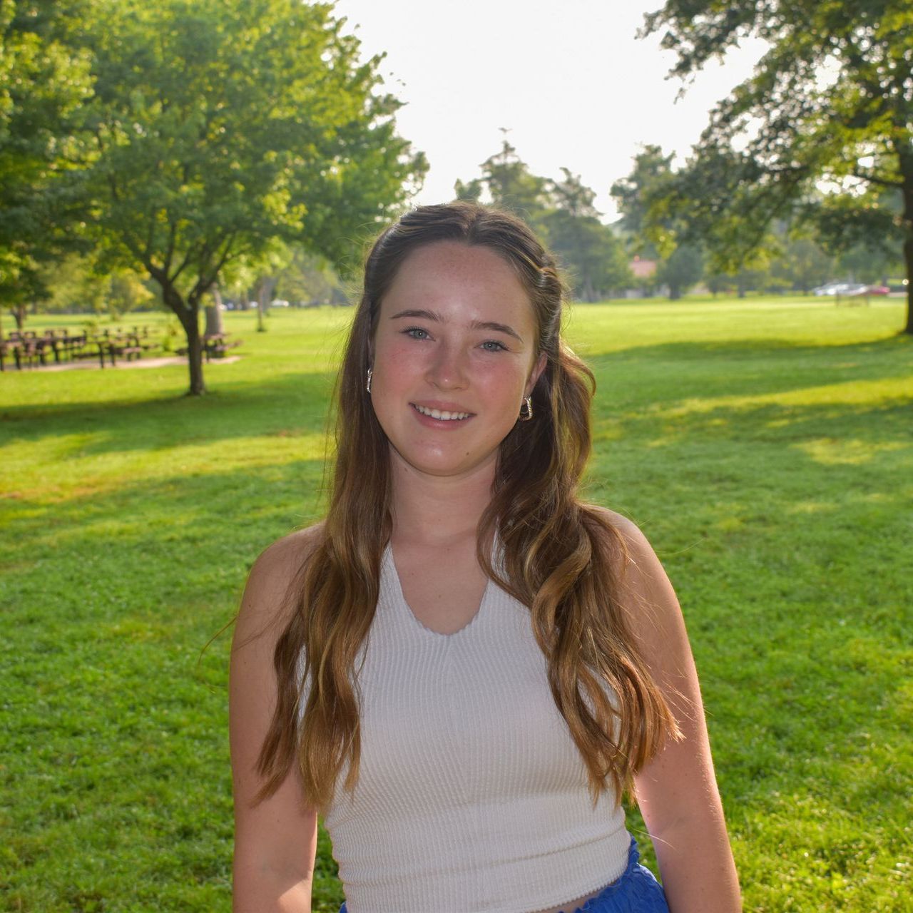 A young woman in a white tank top is sitting in the grass in a park.