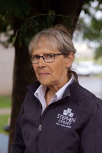 A woman wearing glasses and a jacket is standing in front of a tree.
