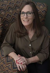 A woman wearing glasses is sitting in a chair with her hands folded.