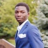 A young man in a blue suit and white shirt is standing in front of trees.