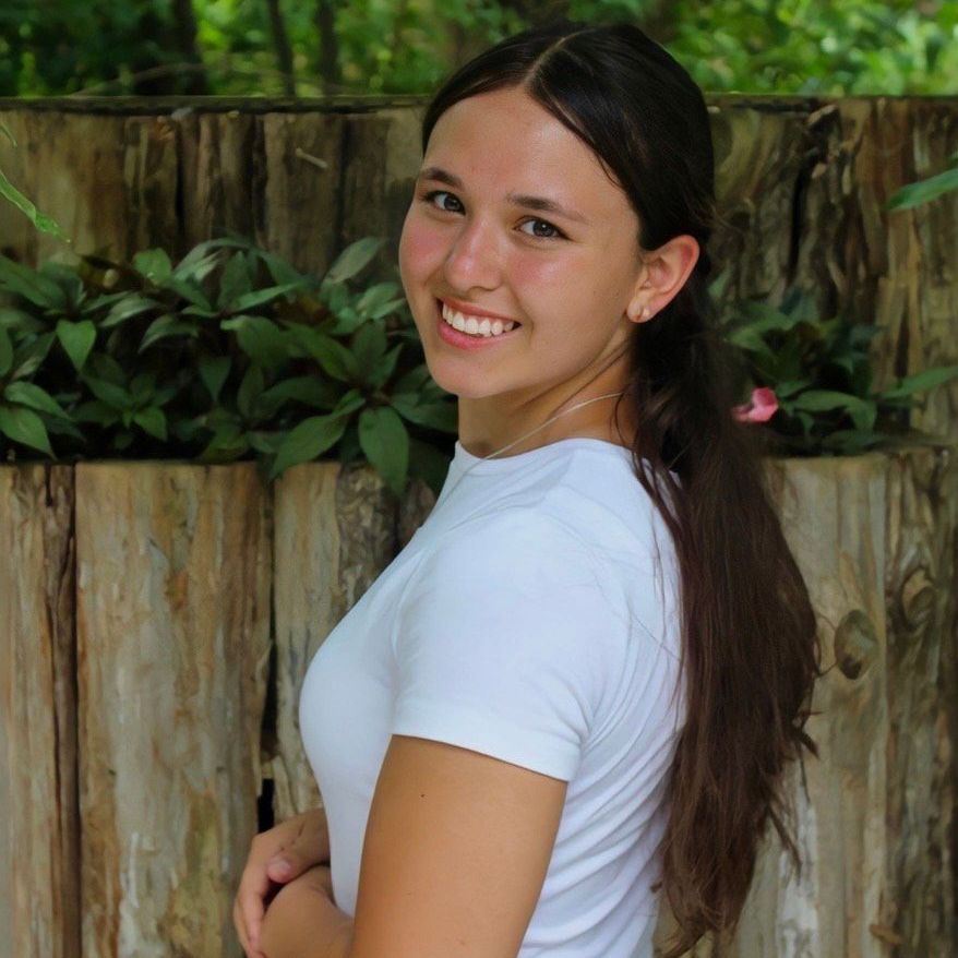 A young woman in a white shirt is smiling in front of a wooden fence.