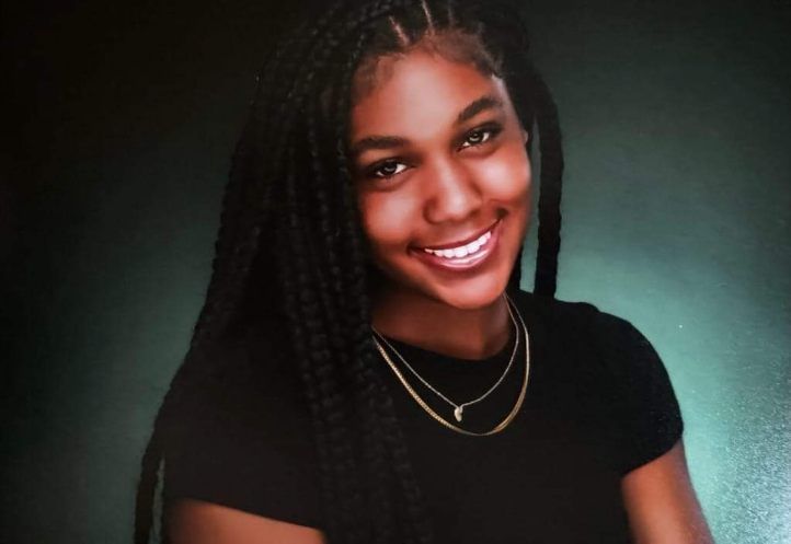 A young woman with braids is smiling for the camera while wearing a black shirt and a gold necklace.