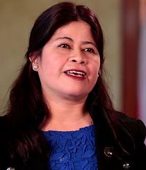 A woman in a blue dress and black jacket is smiling.