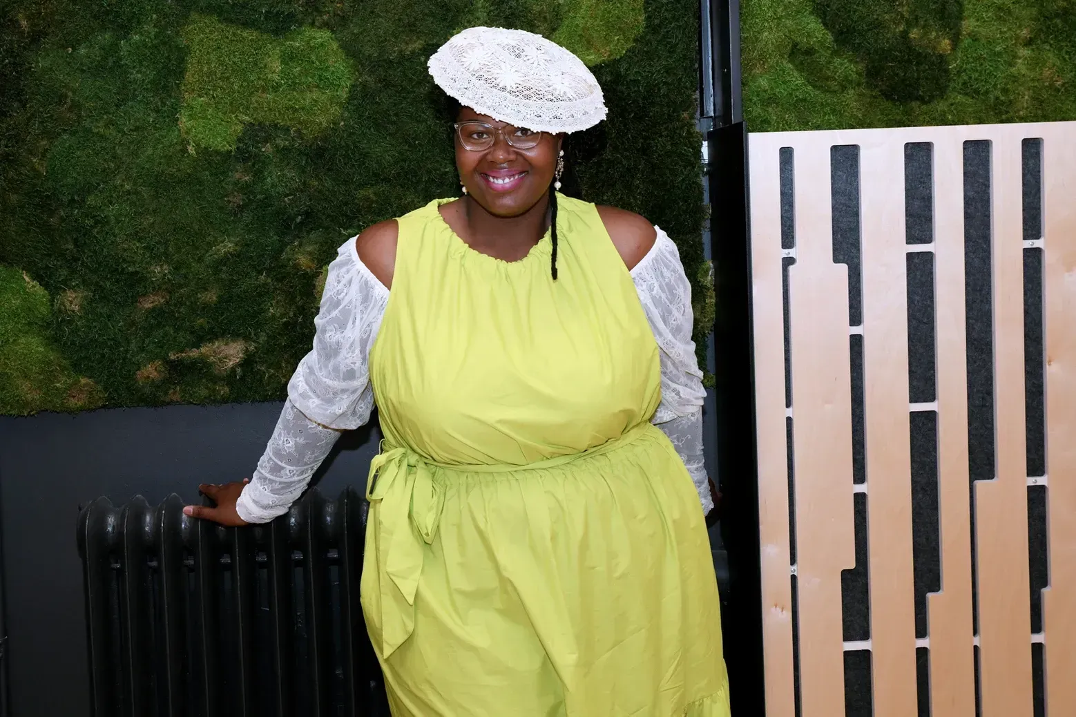A woman in a yellow dress and white hat is standing next to a radiator.