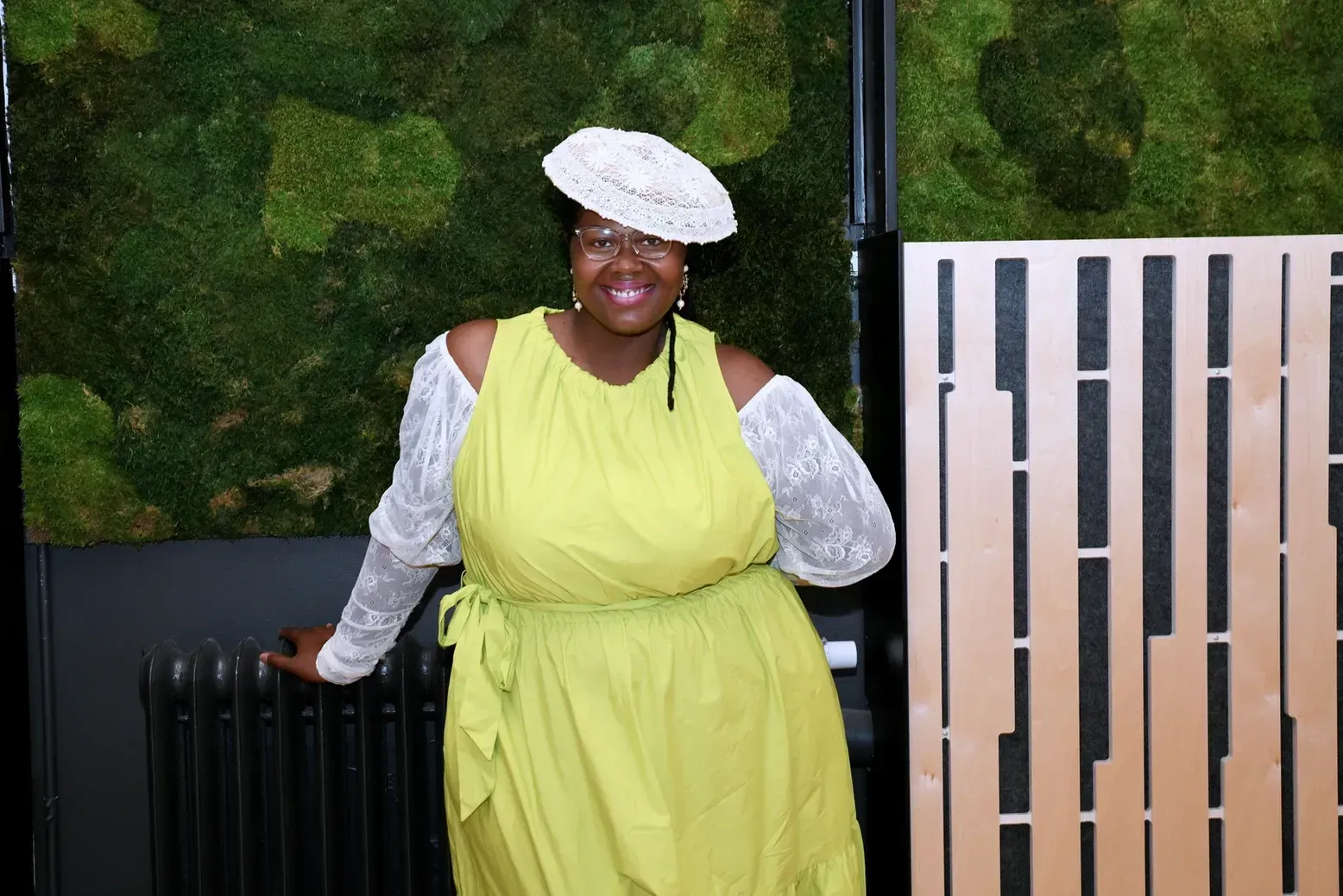 A woman in a yellow dress and white hat is standing in front of a moss wall.