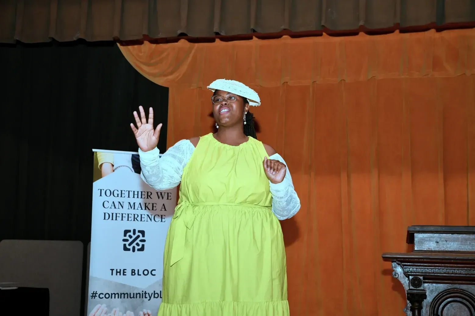 A woman in a yellow dress and white hat is standing on a stage.