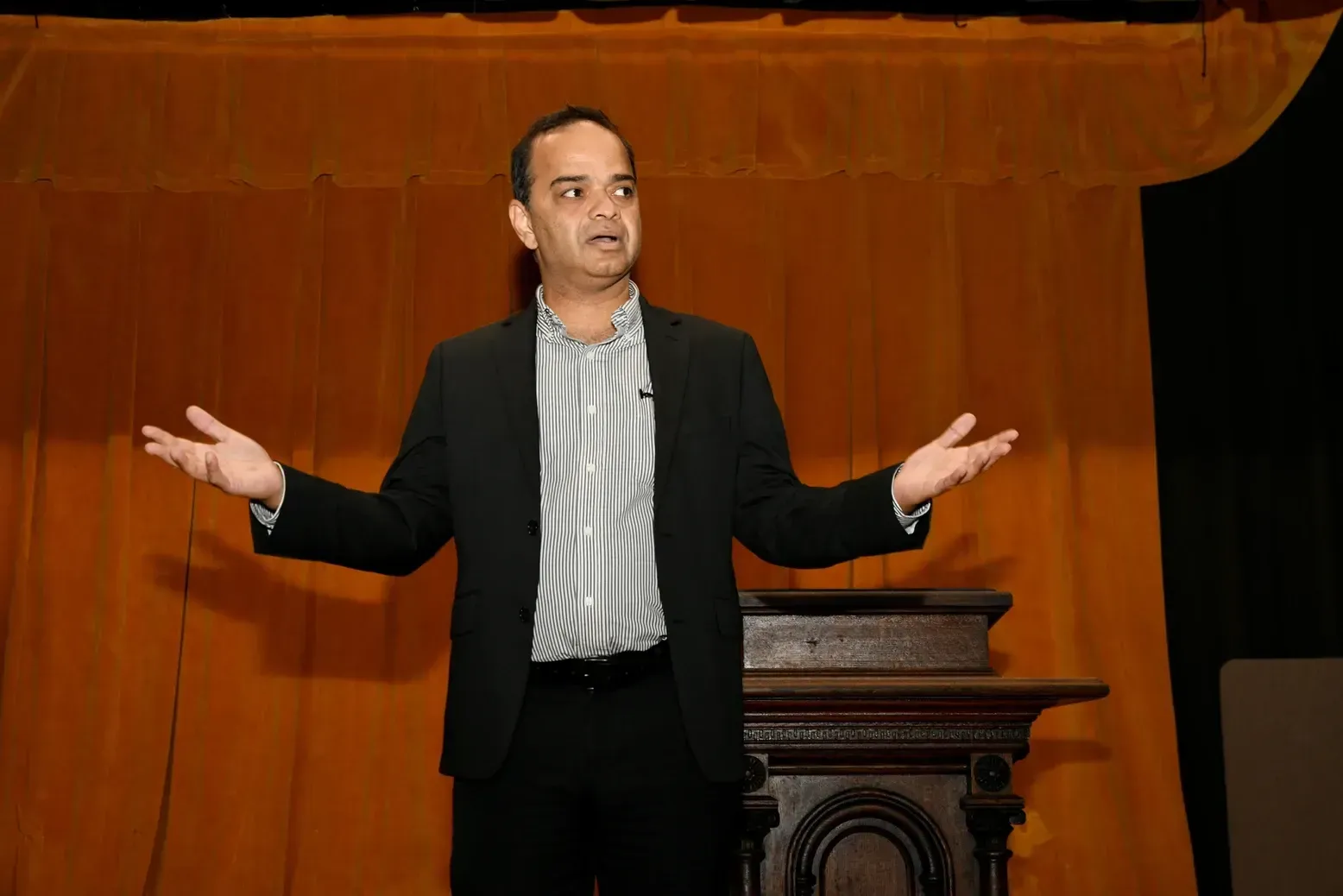 A man in a suit is standing in front of a podium with his arms outstretched.