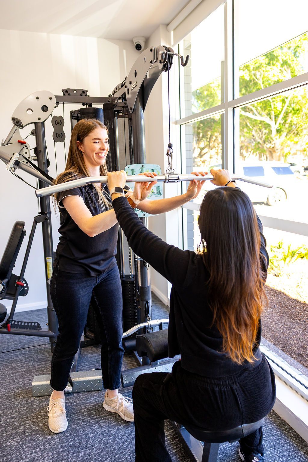 Two women exercising with a cable machine in a bright gym near a large window. — Forerunner Physio In Taree, NSW