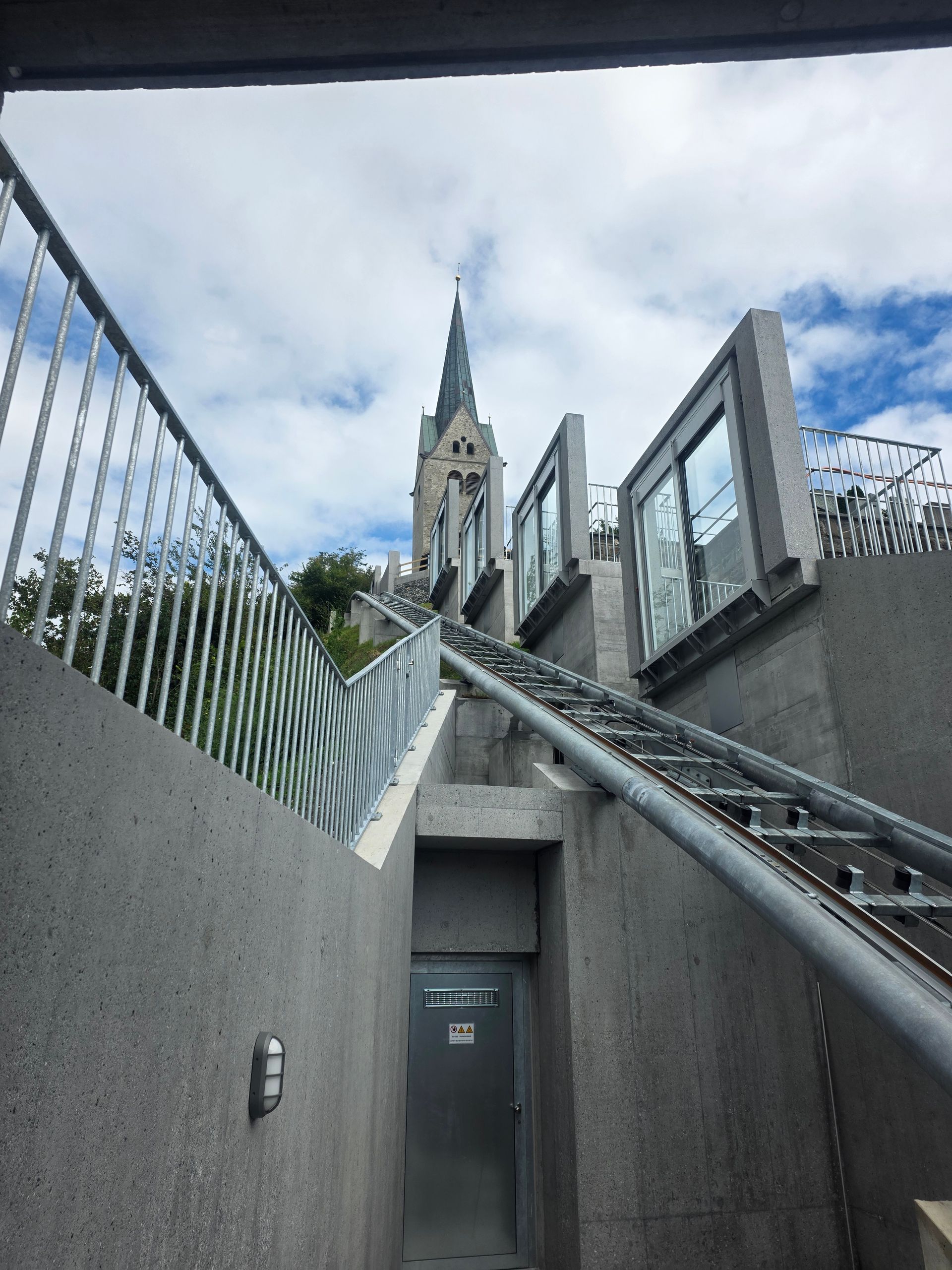 Betontreppen und ein ansteigender Weg führen zu einem Kirchturm. Bewölkter Himmel darüber.