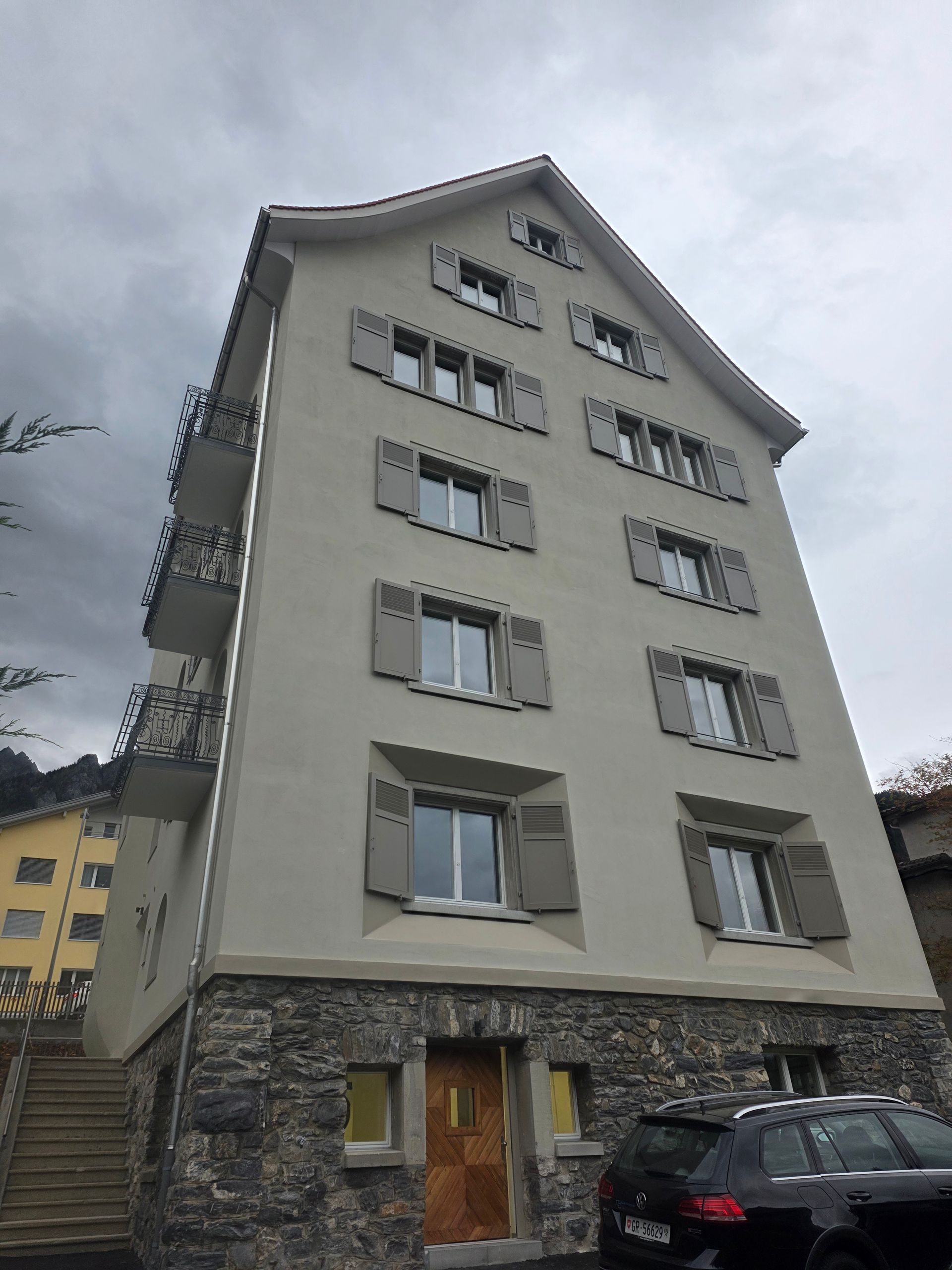 Mehrstöckiges Gebäude mit grauer Fassade, Steinsockel, Fensterläden und Balkonen. Ein Auto parkt davor.