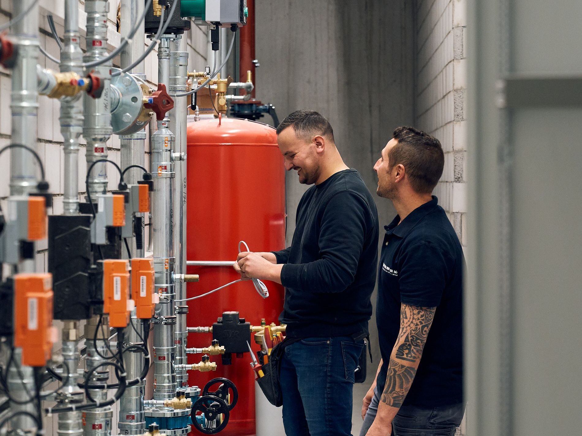 Zwei Techniker arbeiten in einer Industrieanlage an Rohrleitungen; einer hält ein Werkzeug, beide lächeln.