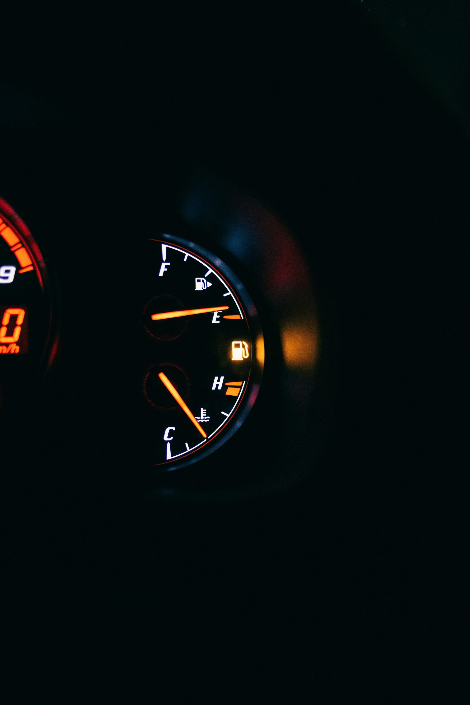 A car's dashboard at night, showing a fuel gauge nearing empty and an illuminated low-fuel warning light.