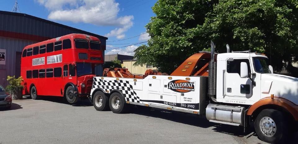 A white tow truck is towing a vintage red double-decker bus in a paved lot under a blue, cloudy sky.