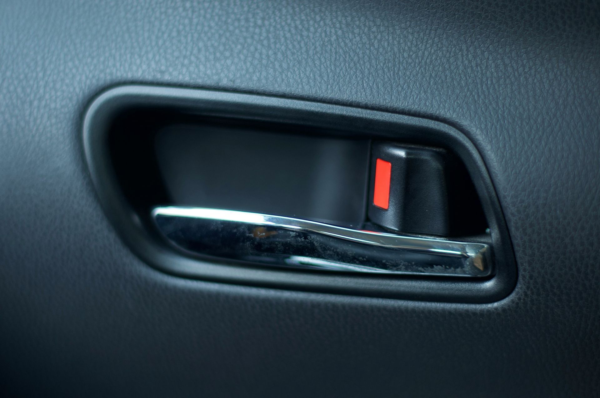 A chrome interior car door handle against a dark gray textured panel, with a small red indicator showing the lock is engaged.
