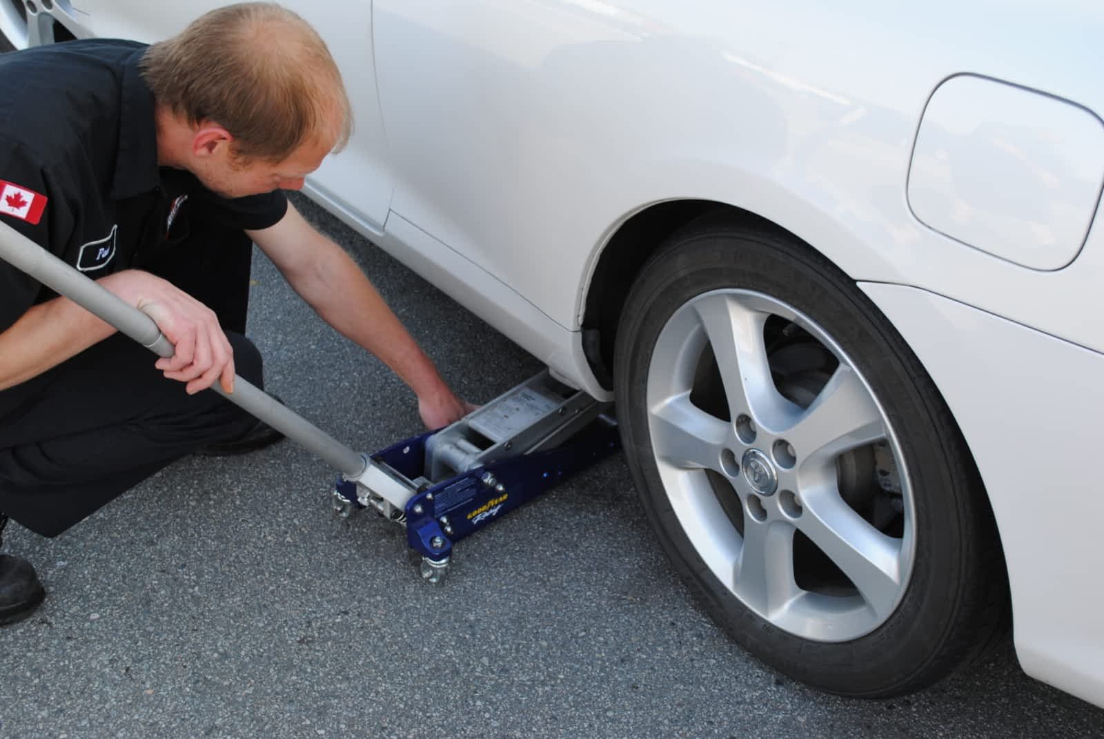 A mechanic positions a blue floor jack under the side of a white car parked on asphalt.