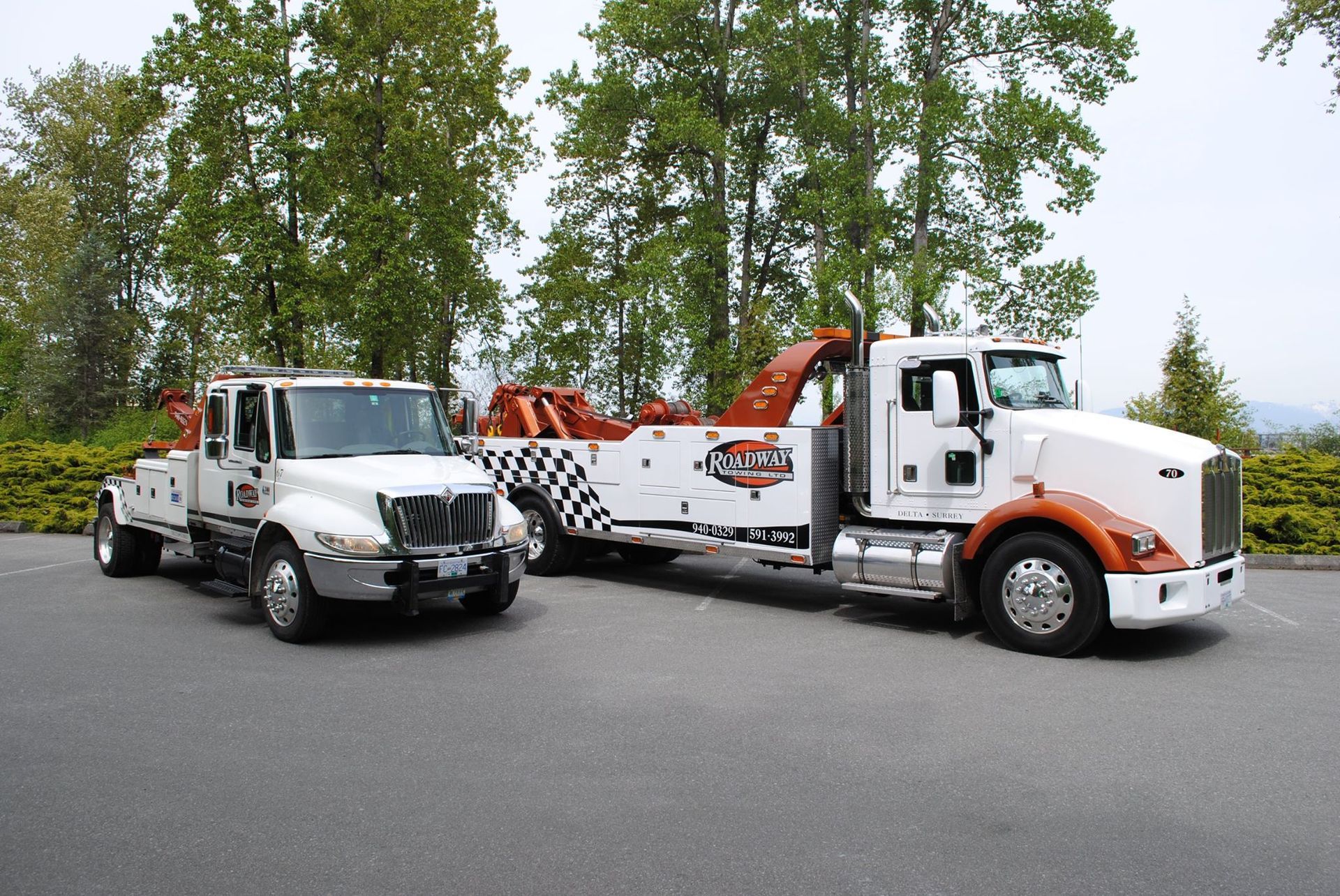Two white heavy-duty tow trucks with orange accents parked side-by-side on an asphalt lot against a backdrop of trees.
