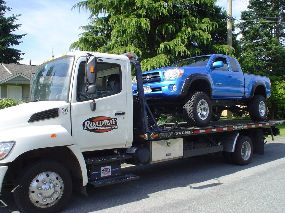 A white tow truck is towing a vintage red double-decker bus in a paved lot under a blue, cloudy sky.