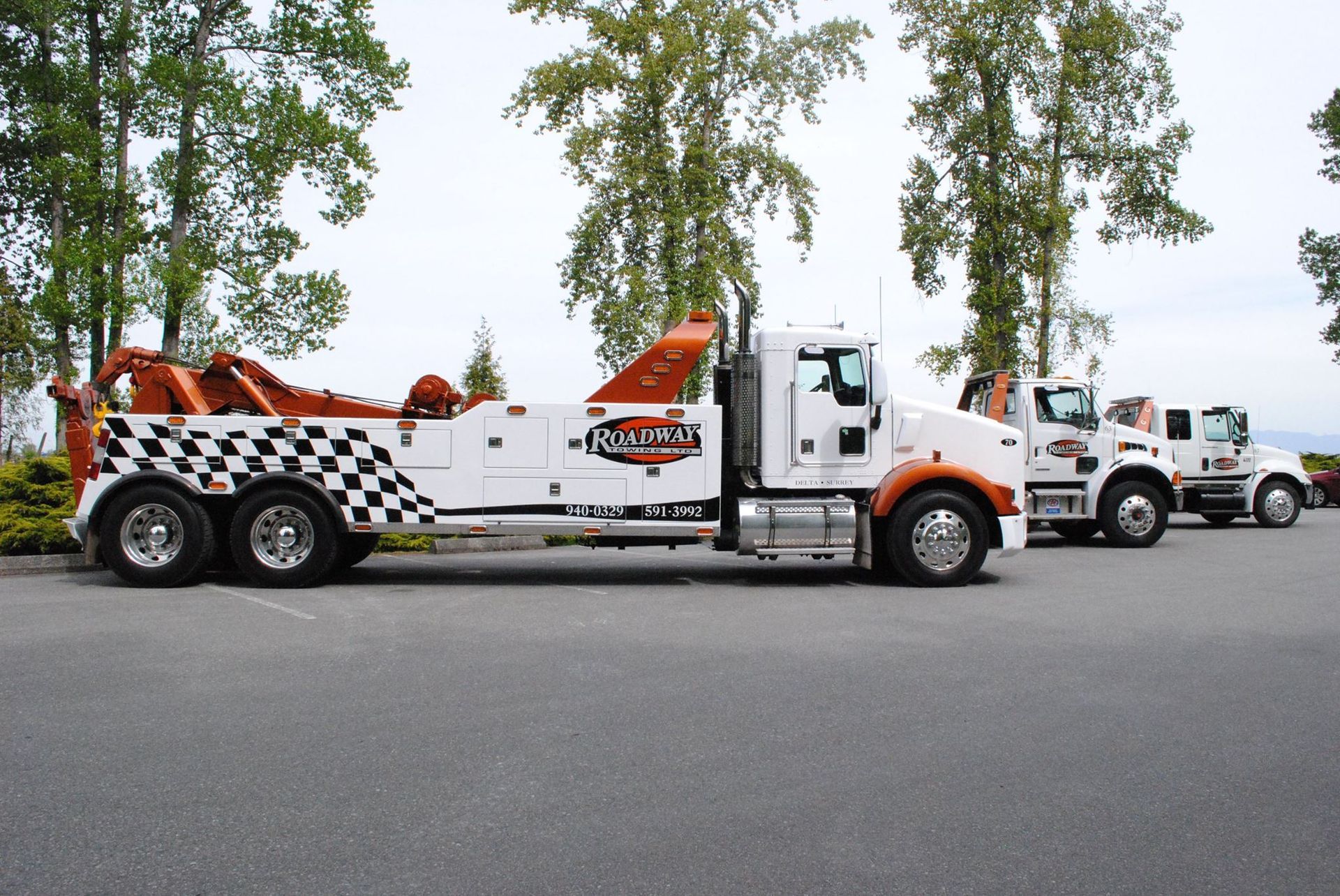 Three white and orange heavy-duty tow trucks parked in a row on a paved lot near trees.