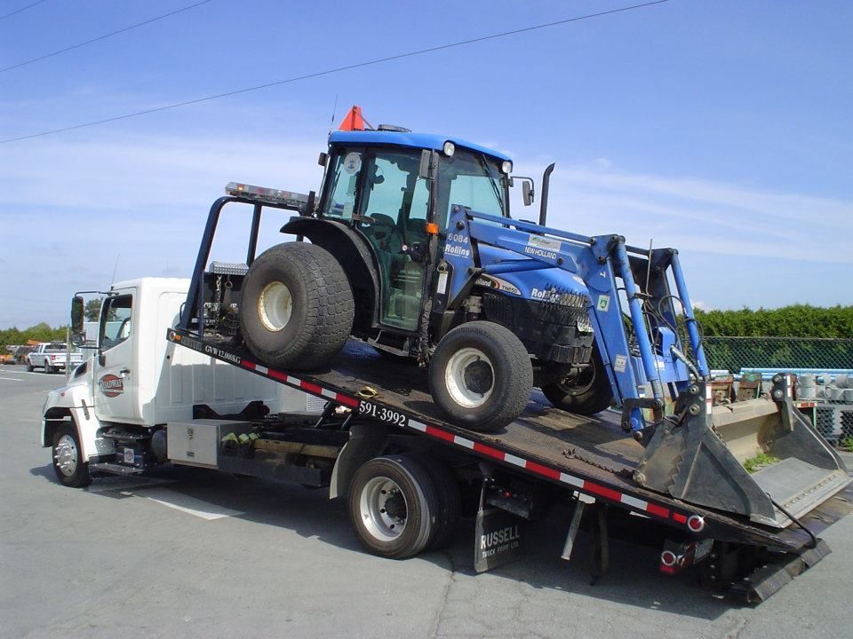 A white flatbed tow truck carries a blue tractor with a front loader on a sunny day.