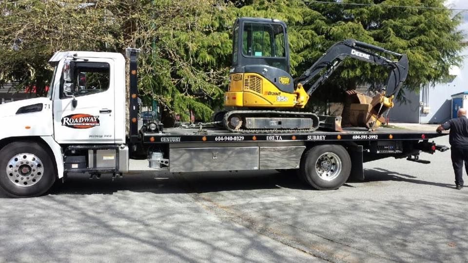 A white flatbed tow truck carries a yellow Deere mini-excavator with a person standing nearby on the right.