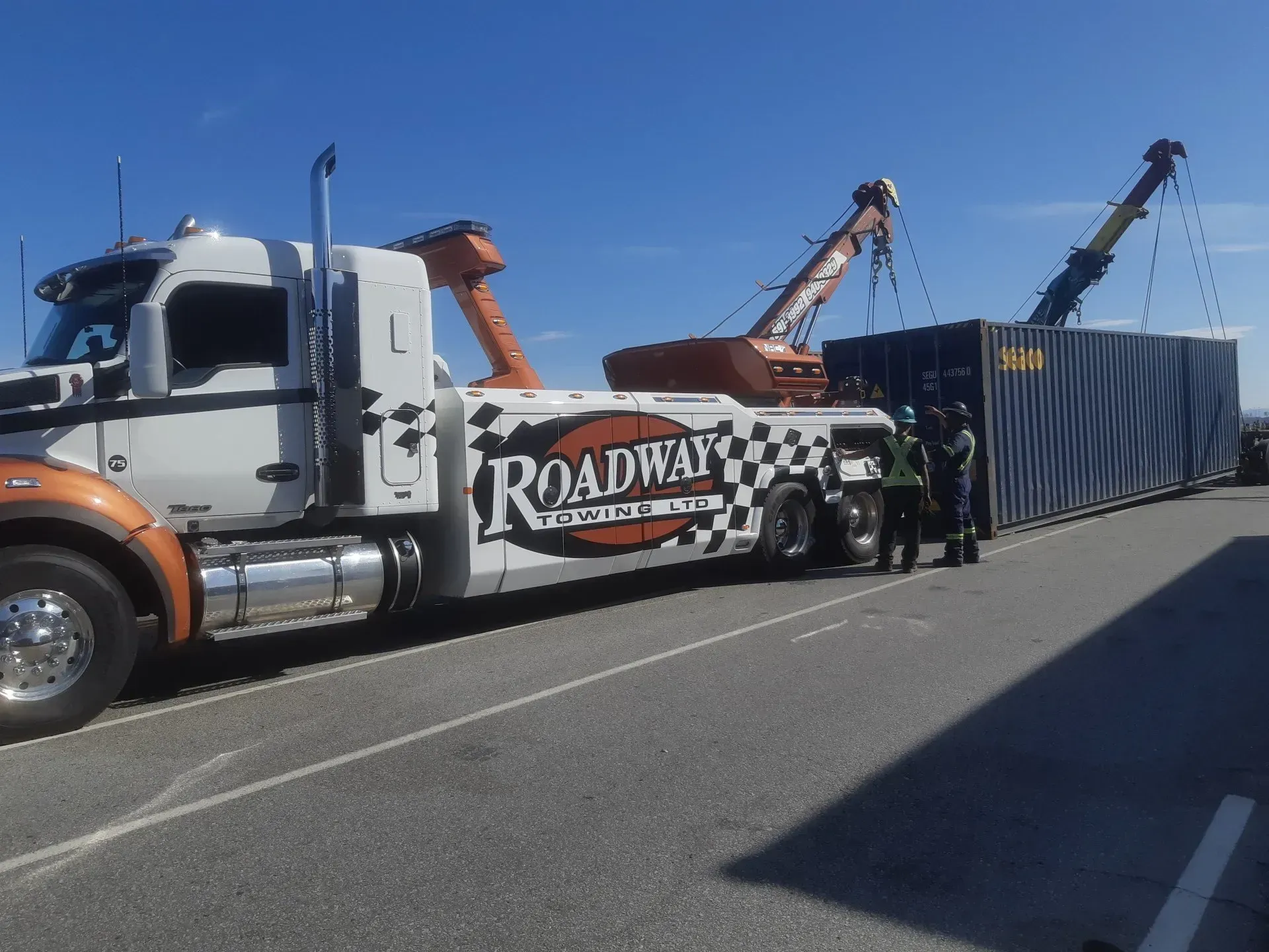 A white tow truck is towing a vintage red double-decker bus in a paved lot under a blue, cloudy sky.