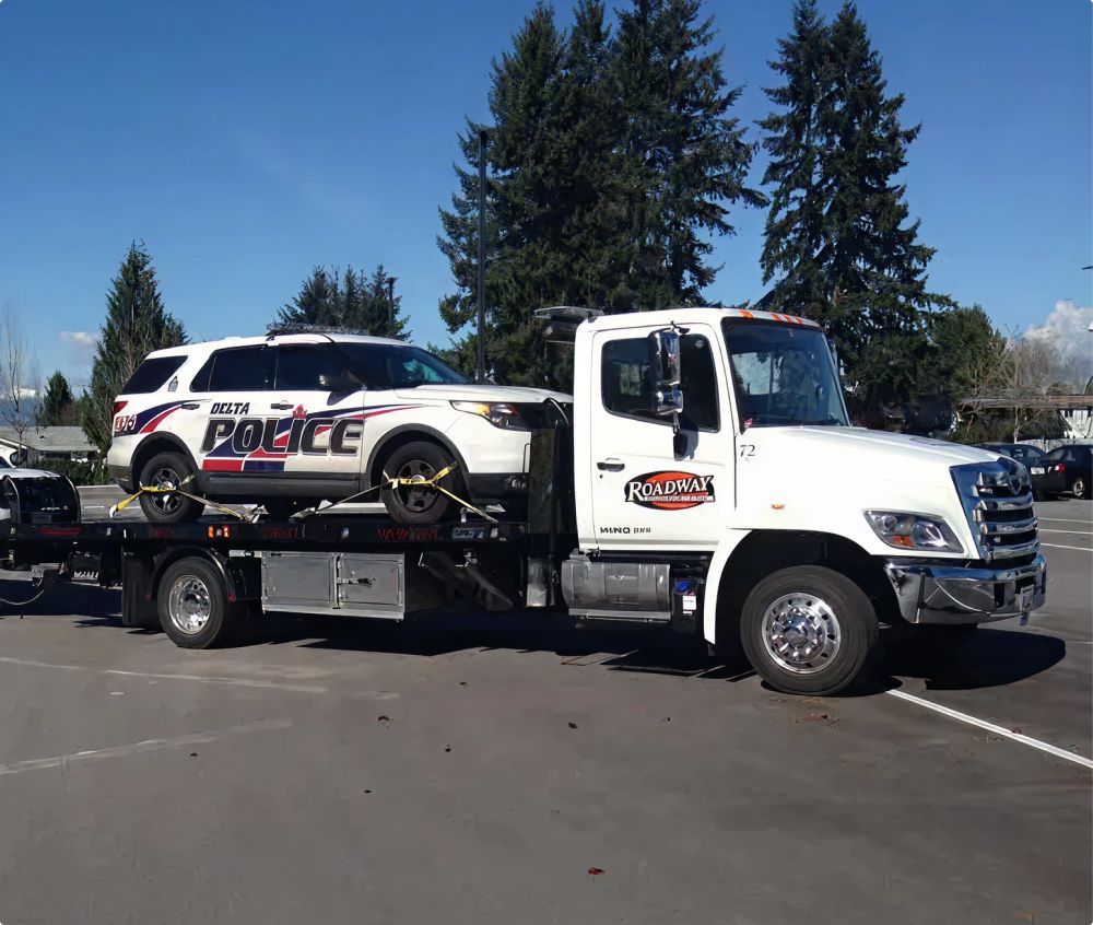 A white tow truck transports a white police SUV in a sunny, paved parking lot.