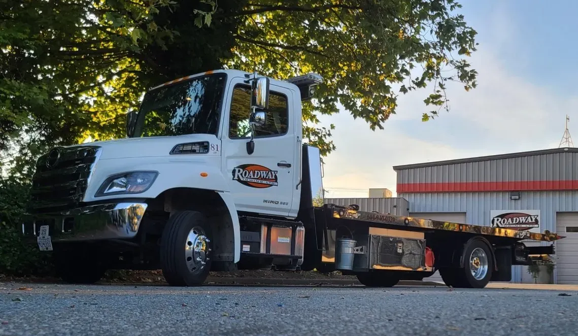 A white Hino tow truck parked on gravel in front of a metal warehouse at dusk, with trees in the background.