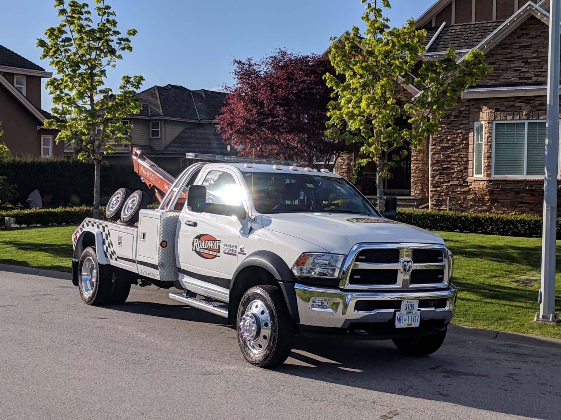 A white Ram tow truck parked on a residential street in front of a house with stone siding and green lawns.