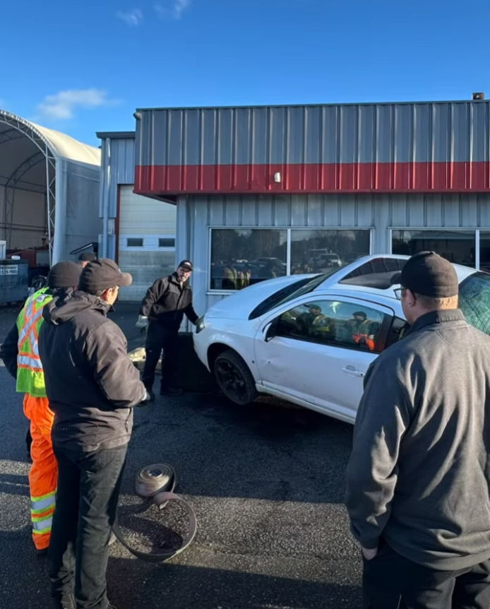 A white car crashed against a building wall while a group of people look on in an outdoor parking area.
