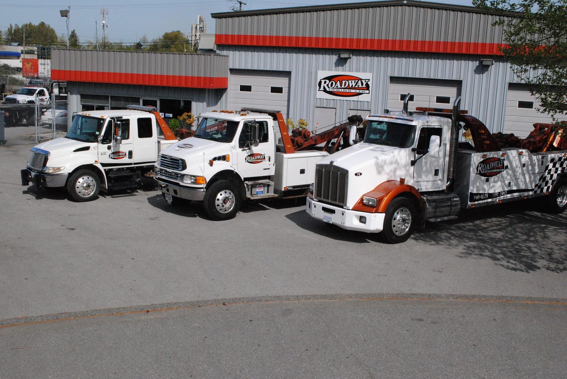 Three white tow trucks parked in front of a gray industrial building with orange trim under a blue sky.