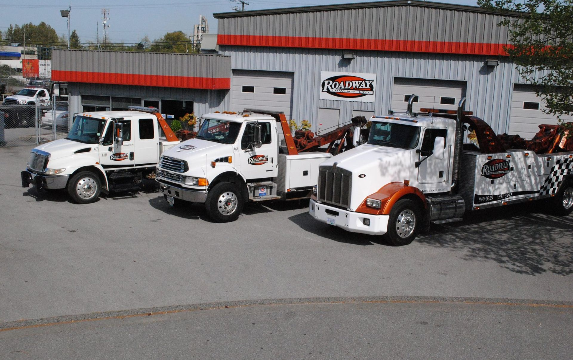 Three white tow trucks parked in front of a light gray industrial building with a red horizontal stripe.