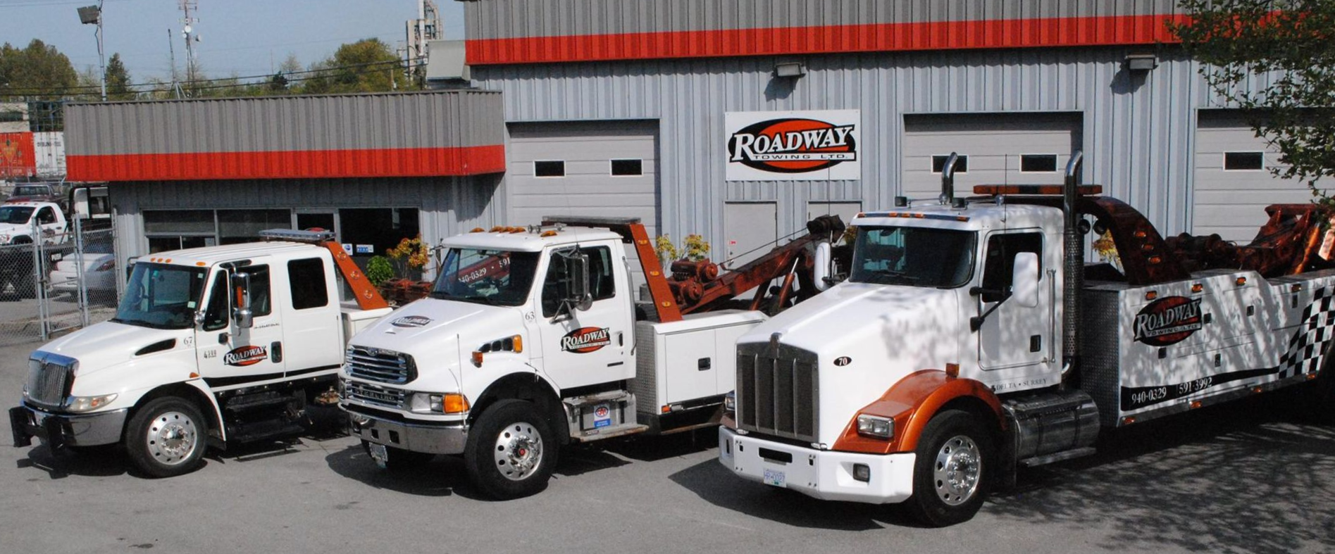 Three white tow trucks parked in front of a grey warehouse building with red trim.