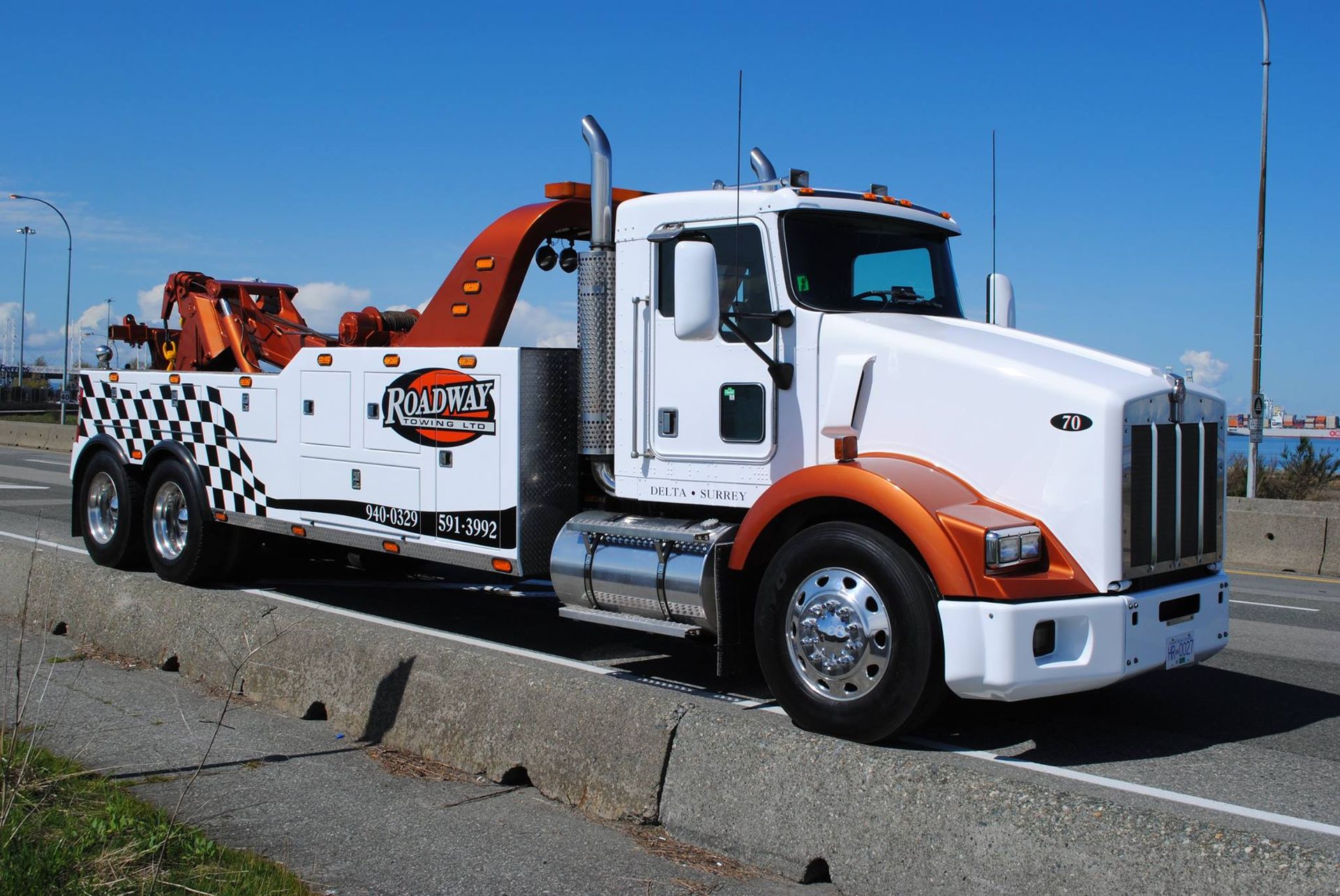 A white and orange tow truck with a checkered pattern on the back parked on the side of a road against a blue sky.