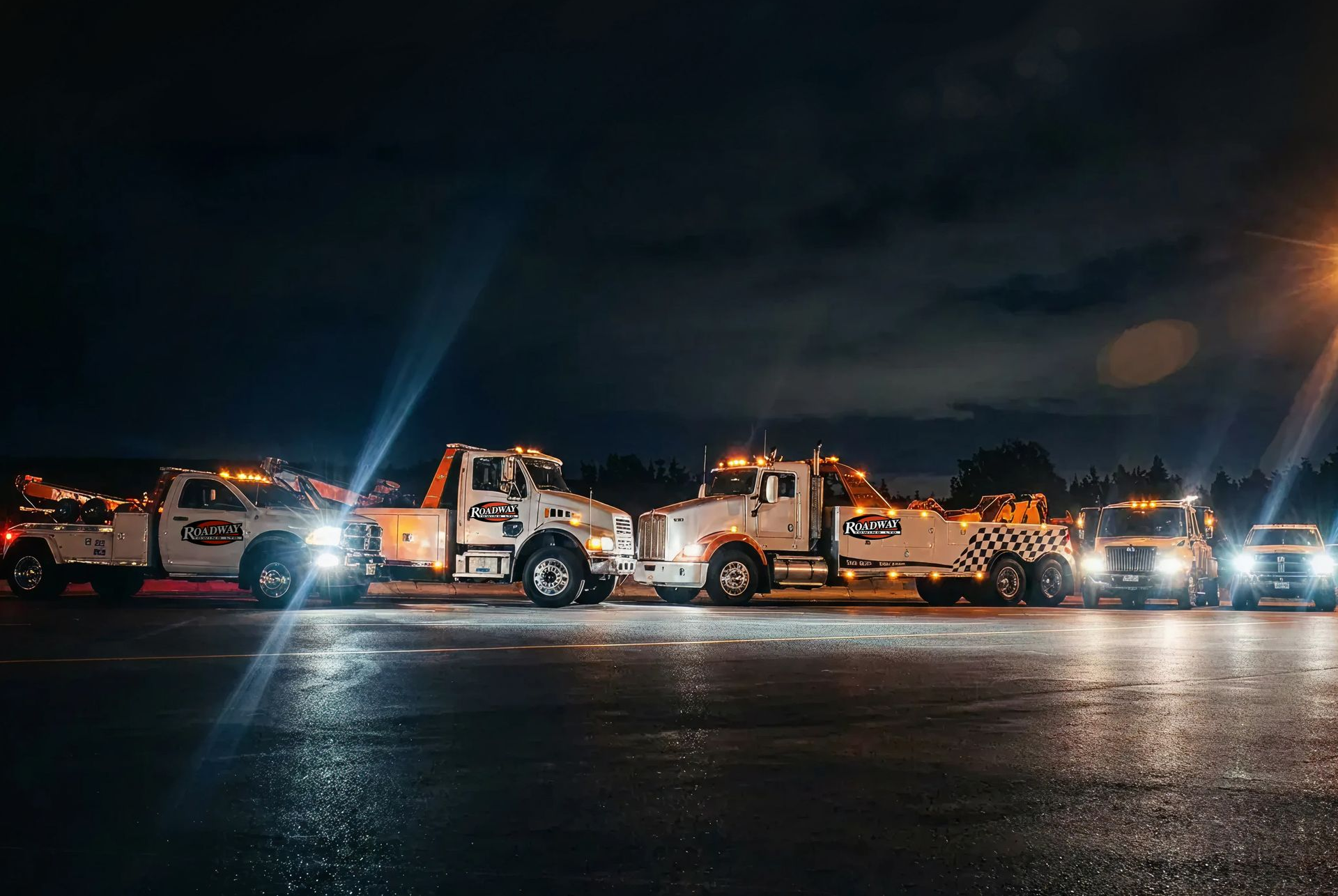 A row of white tow trucks parked in a line at night with their headlights glowing on a dark, wet asphalt lot.