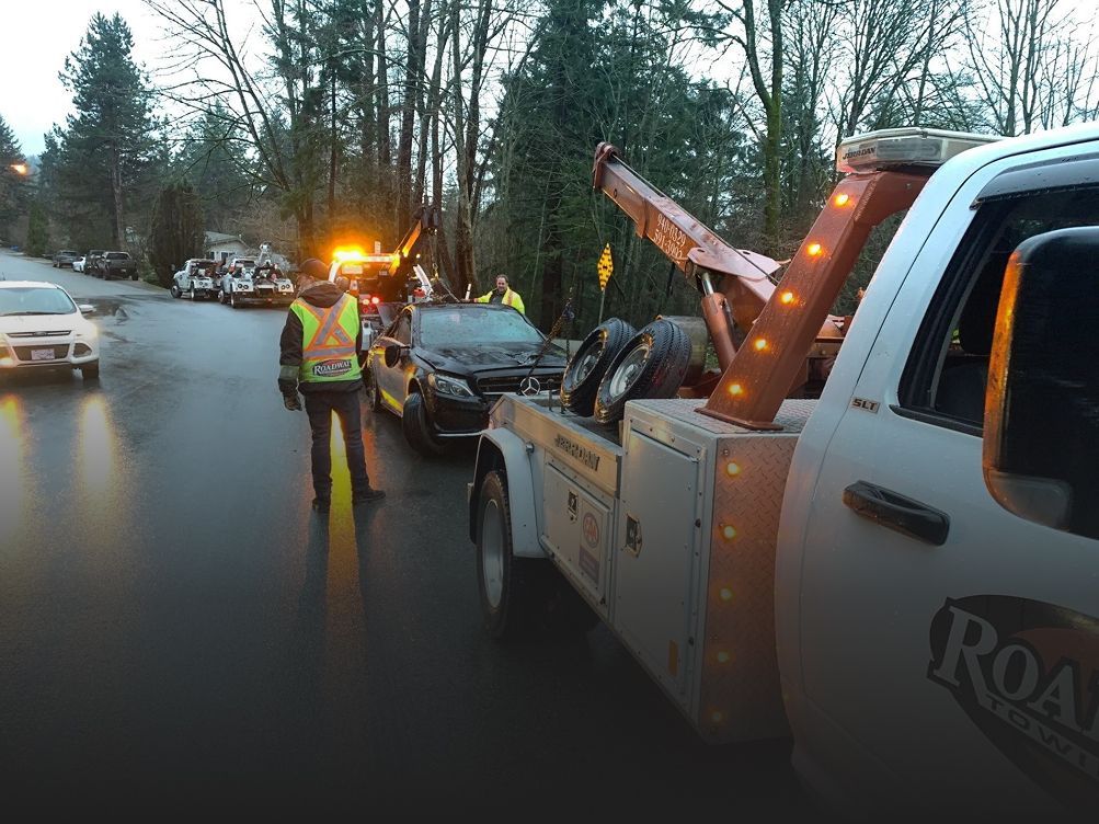 A tow truck driver in a high-visibility vest prepares to load a dark sedan onto a flatbed on a wet, tree-lined road.
