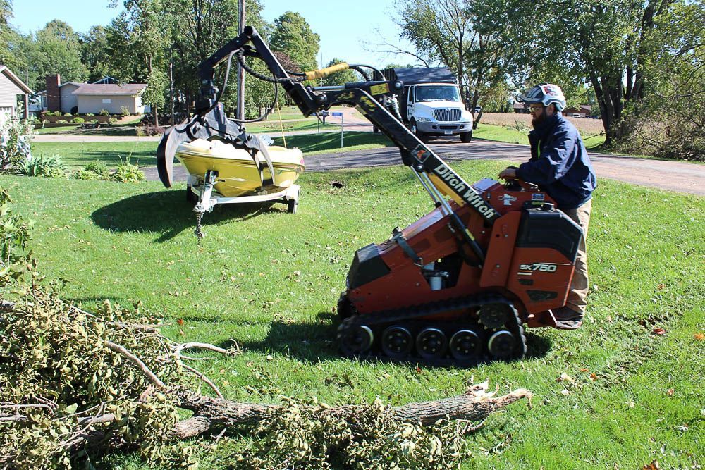 A man is driving a tractor in a yard with a boat in the background.
