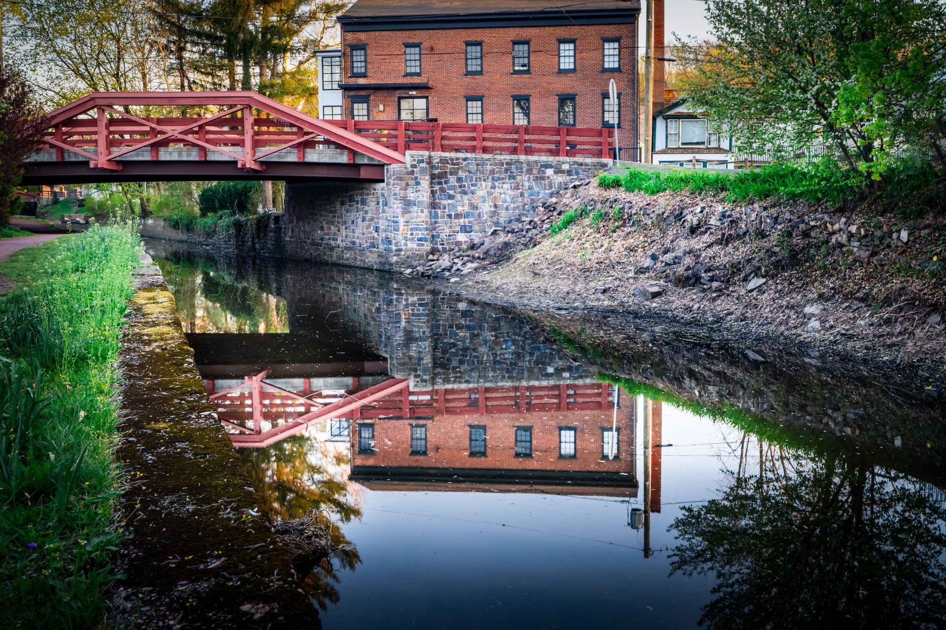 Brick building reflected in canal water beneath a red bridge. Green grass and trees line the canal.